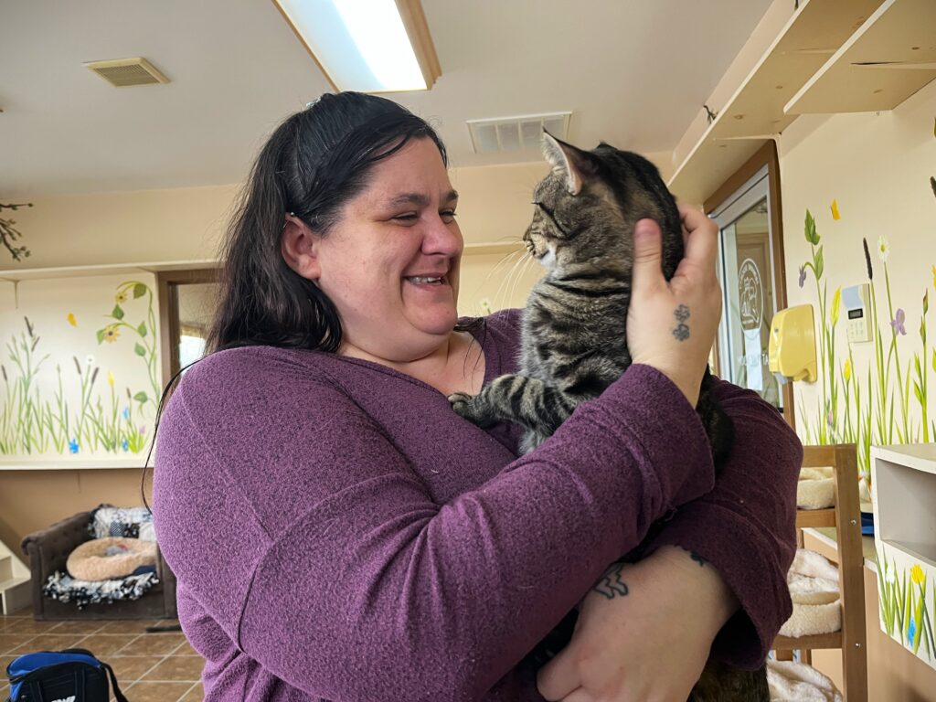 A woman in a purple shirt holds a tabby cat in her arms and smiles at him. The cat looks back at her inquisitvely.