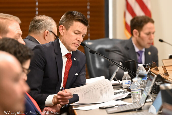 A man wearing a dark blue suit over a white shirt and red tie turns a page. He is seated at a curving desk with other men in suits behind and in front of him. In the far background wooden venetian blinds can be seen.
