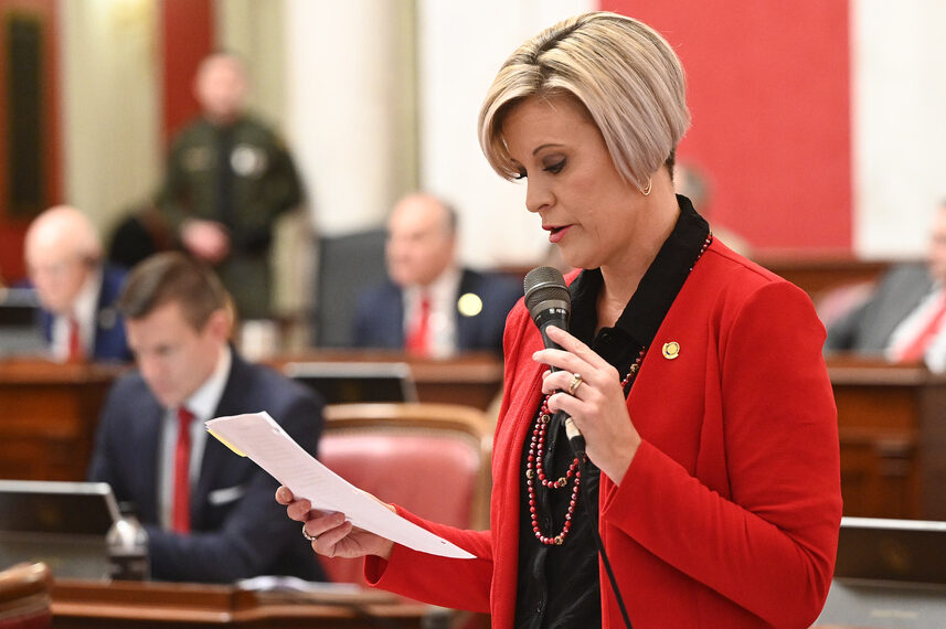 A woman in formal attire stands before several wooden desks and speaks into a microphone. She is looking down at a piece of paper in her hand.