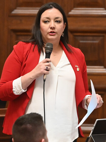 A woman in formal attire stands up at a wooden desk and speaks into a microphone. She is holding a piece of paper in her hand.