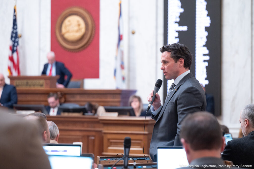 A man in a suit and tie stands up and speaks into a microphone. He is surrounded by people working at wooden desks, and behind him the seal of the West Virginia House of Delegates, the West Virginia and American flags, and a black-and-white screen are visible.