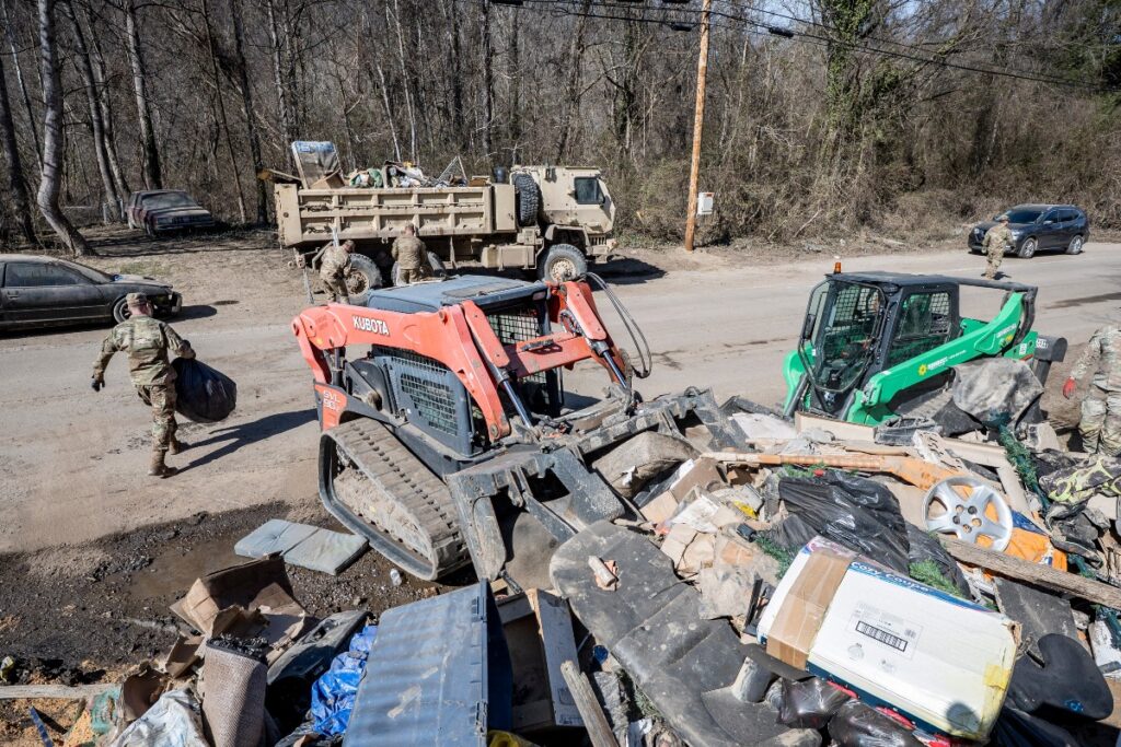 two small skid steer loaders pile damaged property on the side of a road. Around them individuals in fatigues carry bags to a military hauling truck staged in front of a wooded lot.