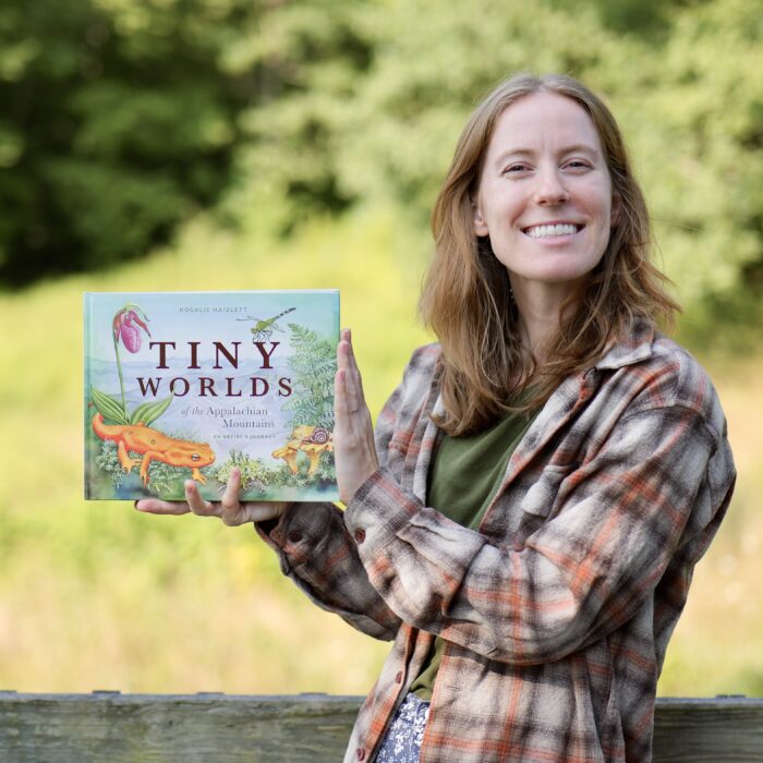 White woman outside in front of trees holding a book that reads, "Tiny Worlds" on the cover.