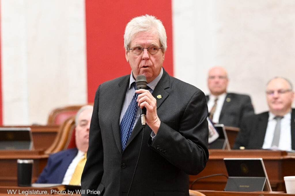 A man with white hair speaks into a microphone he is holding in his left hand wears a black suit over a light blue shirt and a blue tie. Behind him other men wearing suits sit at wooden desks in front of a white marble wall with cloth accents.