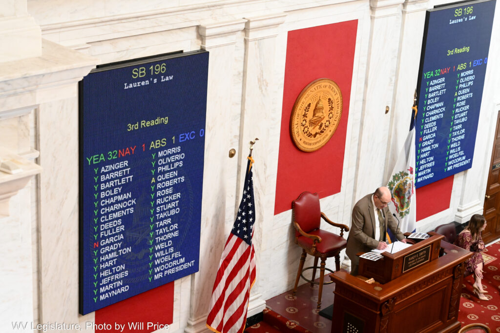 A man wearing a grey suit is seen standing at a podium in a Senate Chamber with marble walls. Two screens show the results of a legislative vote on a bill.