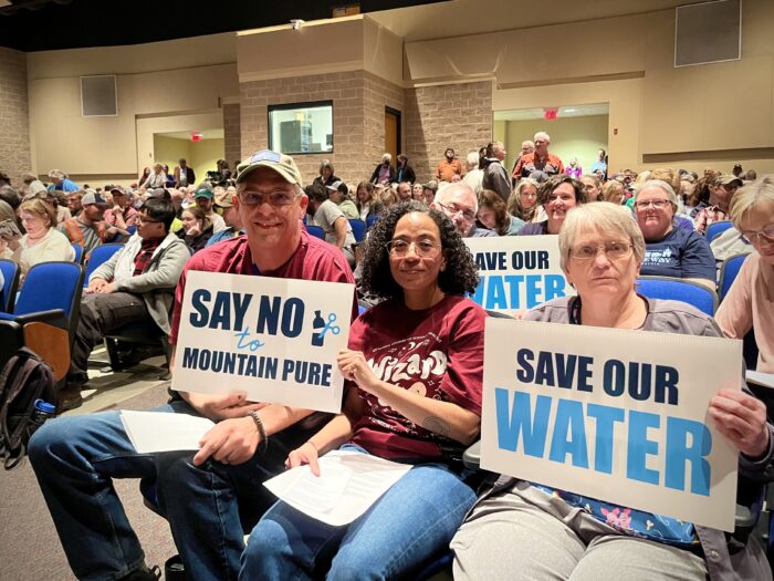 In an auditorium filled with rows of people, three seated up front hold signs that read "Say No To Mountain Pure" and "Save Our Water."