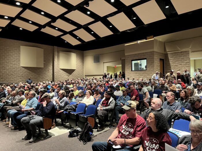 Hundreds of people crowd into an auditorium with blue chairs and cement brick walls.