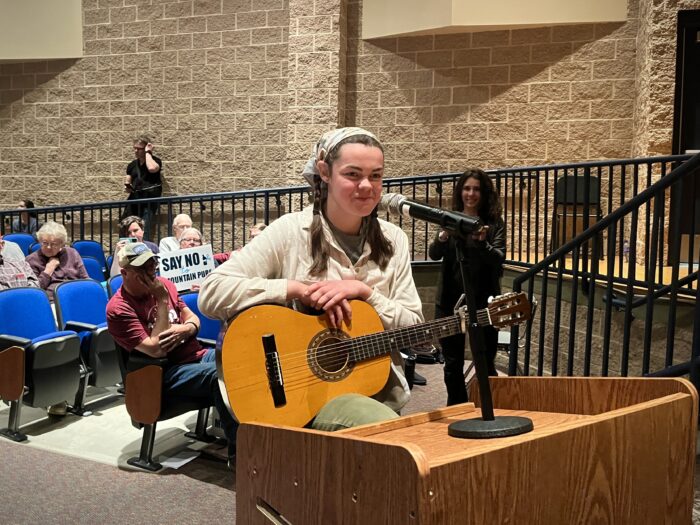 At the front of an auditorium, a woman stands before a podium with a microphone and holds a guitar.