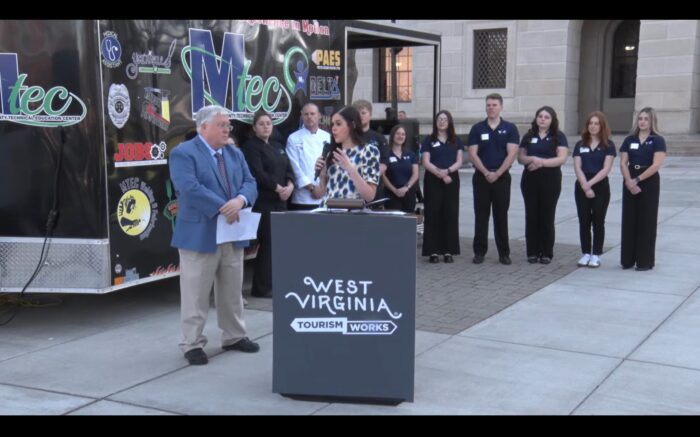 A crowd of people stand around a food truck and a podium. A woman with black hair wearing a polka dot shirt is speaking from the podium to a man wearing a blue suit.