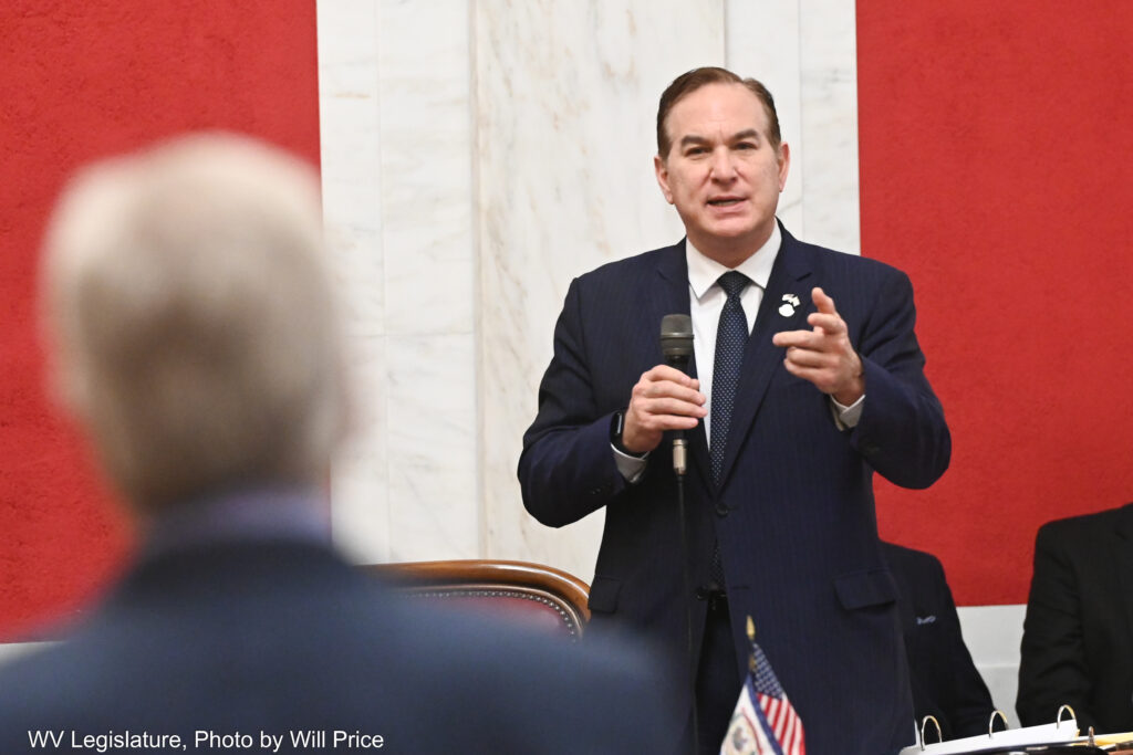 A man wearing a black suit points while speaking into a microphone at another suit-clad man we see in the foreground of the photo. The walls are white marble with red paneling.