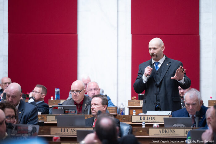 A man in a suit and tie stands up at a desk with a name placard reading "Dillard" and speaks into a microphone, gesturing with his left arm. Around him, other people dressed in formal attire sit at desks with name placards. Behind him, a white marble wall is covered in parts with red velvet.
