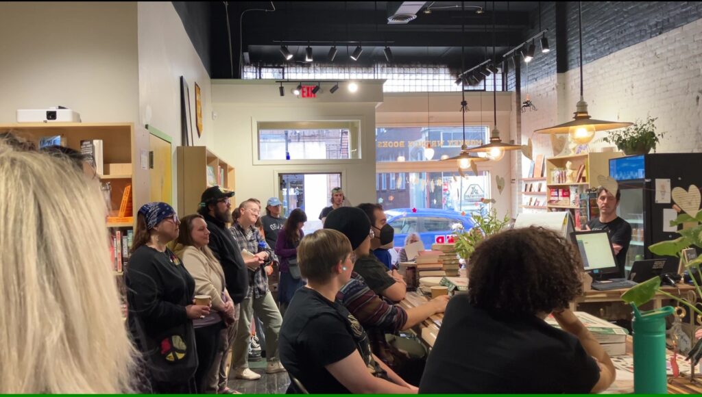 A crowd is arrayed in two parallel lines below a high ceiling. To the right of frame lights hang above a bar where people are seated. To the left of frame some people in the line dip into the alcove of a book shelf. Parked cars and light can be seen through a large window and casement windows in the background, the front of the store.