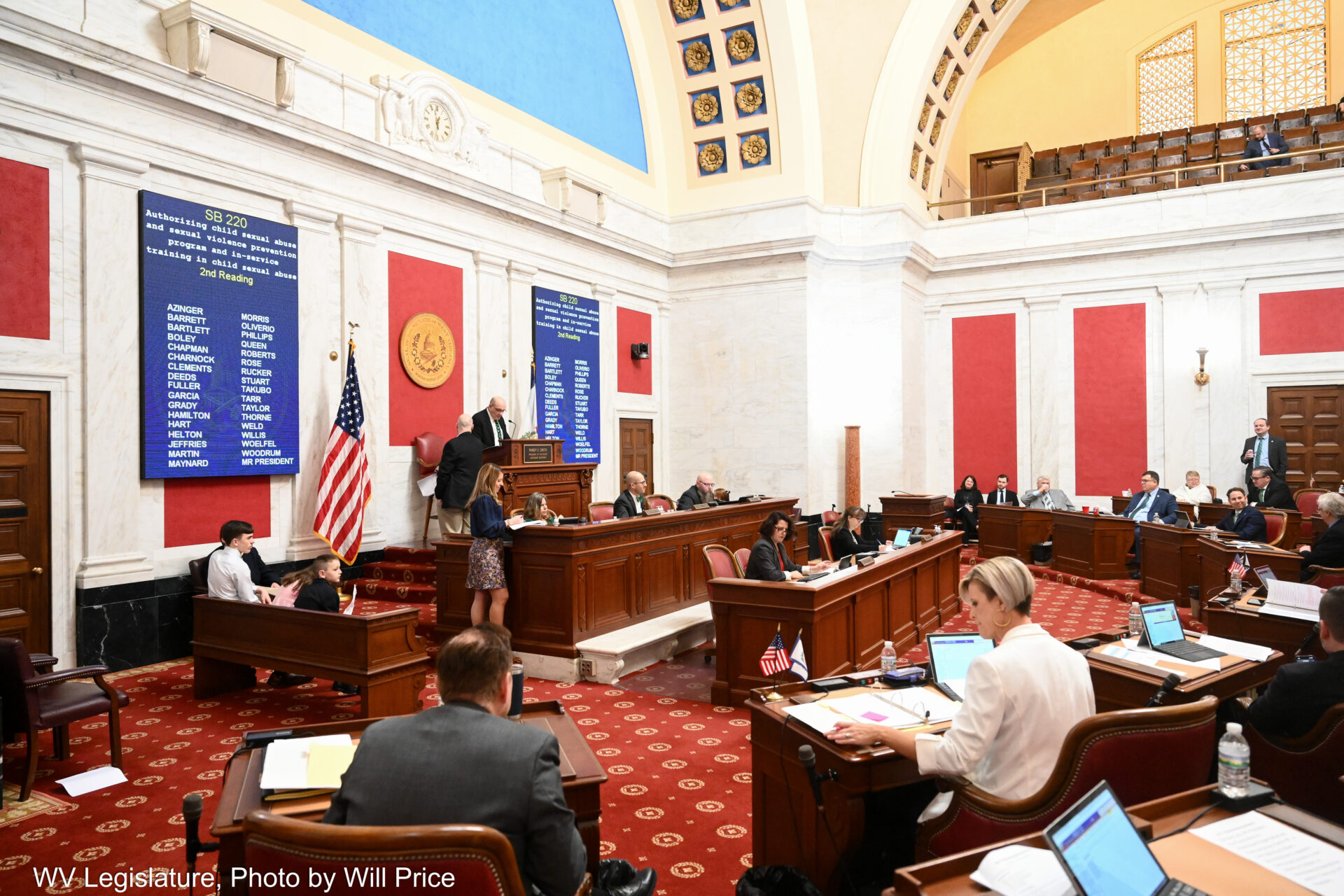 A mid shot of a legislative chamber shows people seated at dark wooden desks on a dark red rug. The image shows a large dais at the front of the room with large screens flanking a man standing above the rest. The screens display a list of names with "Senate Bill 220" at the top. The walls are white marble adorned with red cloth panel accents. In the center background can be seen a large column reaching up to vaulted ceilings out of frame.