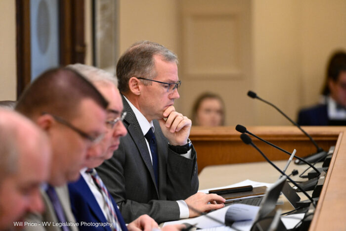 A man wearing glasses and a dark grey suit over a white oxford shirt sits at the end of a row of men in suits with his chin in his hand in a pensive position looking at a screen just out of frame right.