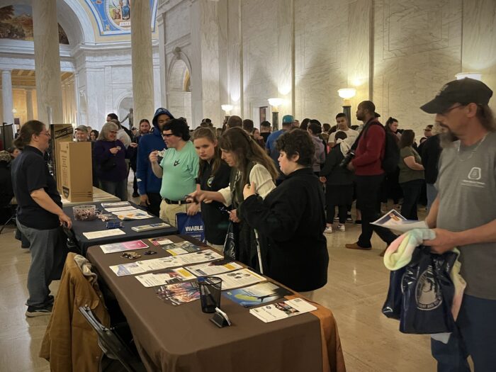 In a marble hallway, a crowd of people stand in front of a row of tables displaying posters, merchandise and informational materials. A couple of people stand behind the tables as if staffing them.