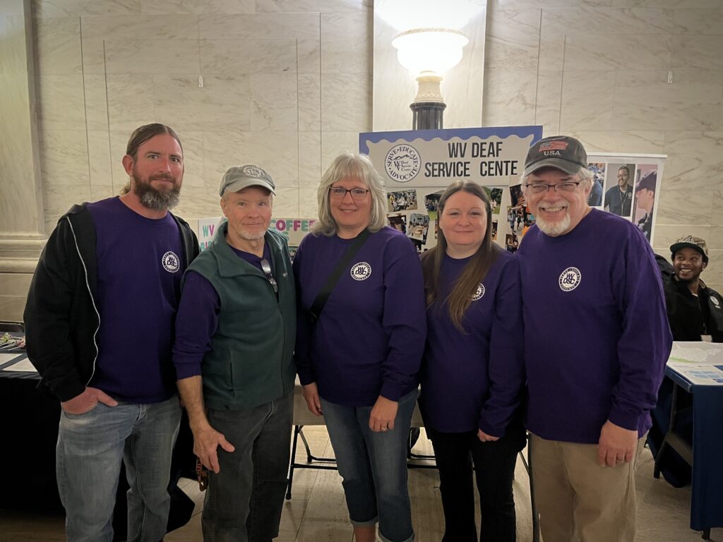 Five people in purple shirts stand together in a group in a marble hallway and smile at the camera. Behind, them a sign on a table reads "West Virginia Deaf Service Center."