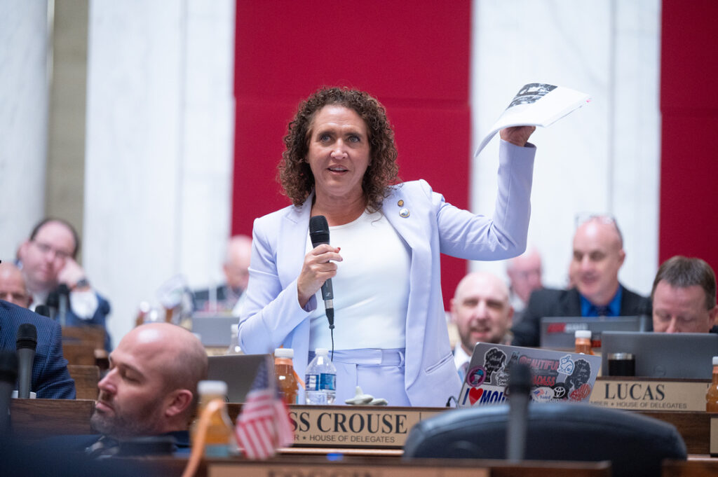 A woman in a light blue pantsuit with a white shirt stands up behind a wooden desk. She holds a microphone in her right hand and a paper packet in her left. Around her, people in formal attire sit at wooden desks with metallic name placards.