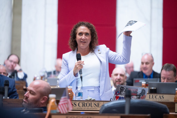 A woman in a light blue pantsuit with a white shirt stands up behind a wooden desk. She holds a microphone in her right hand and a paper packet in her left. Around her, people in formal attire sit at wooden desks with metallic name placards.