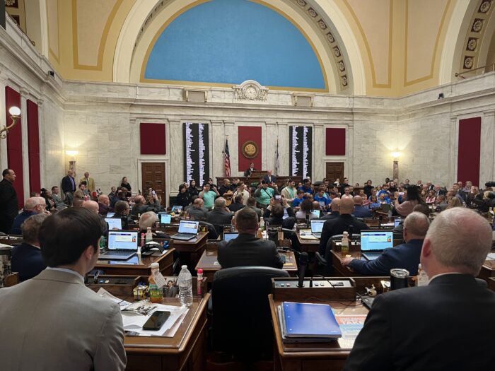 A group of young people stand at the front of the House chamber, a marble room with red carpeting and rows of hardwood desks where delegates in formal attire are seated.