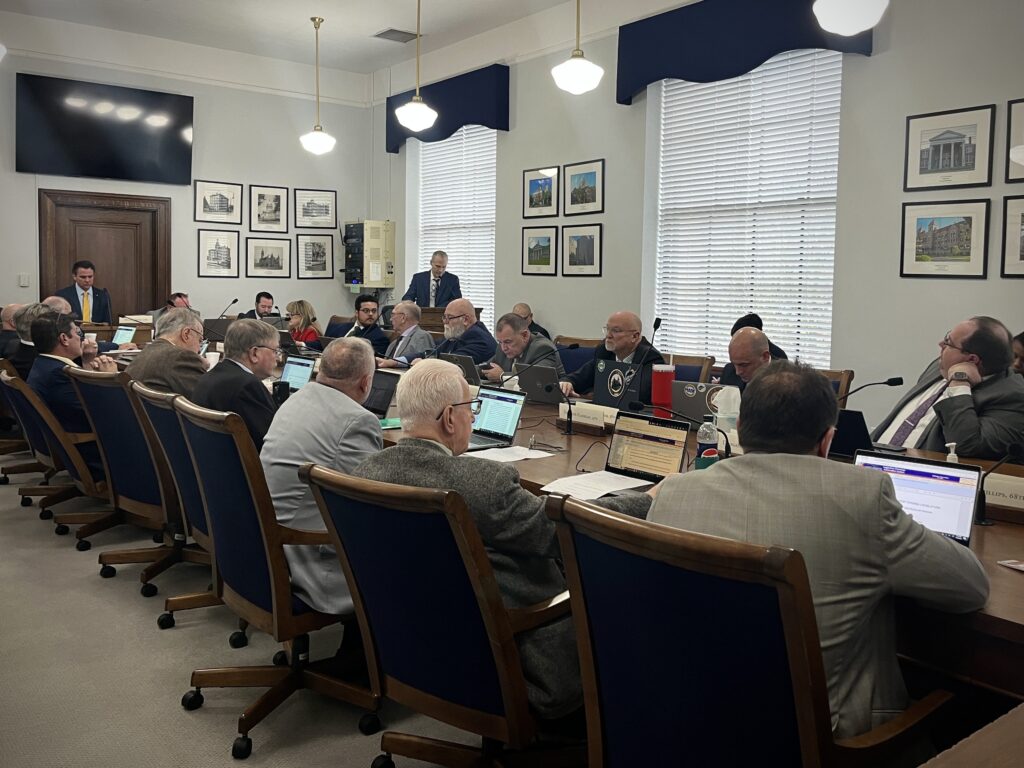 People dressed in formal attire sit around a long, wooden table with laptops out on top of it. At the front of the committee room, a man stands at a podium with and speaks into a microphone.