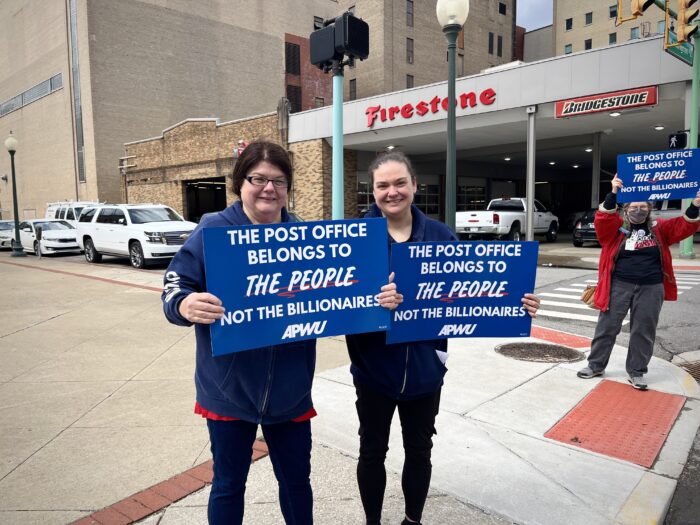 Two women stand on the corner of a city street, smiling toward the camera. They hold up blue signs that read: "The post office belongs to the people, not the billionaires."