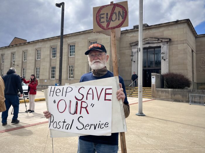 A man in a baseball hat stands outside a concrete building with a tall, glass-paned doorway. He is holding up a handwritten sign that reads "Help Save 'Our' Postal Service," and another small sign on a picket that reads "Elon" with the text crossed out.