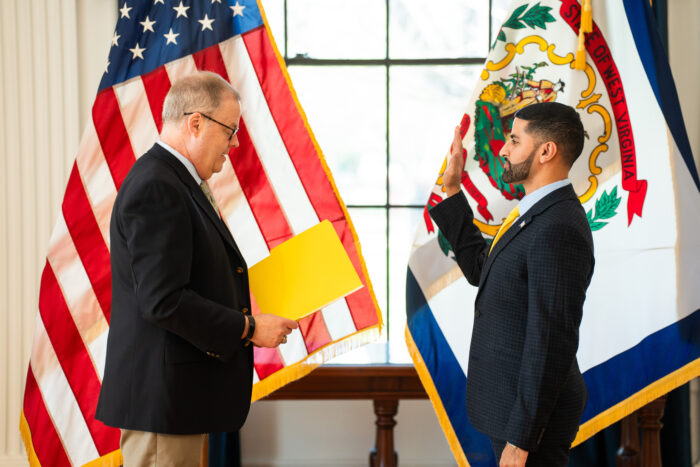 Two men wearing suits stand in front of the U.S. and West Virginia flags. One holds a paper he reads from as the other holds up his right hand as a sign of his oath. The man on the left has grey hair and wears glasses. The man on the right has dark hair and a closely shaved beard.