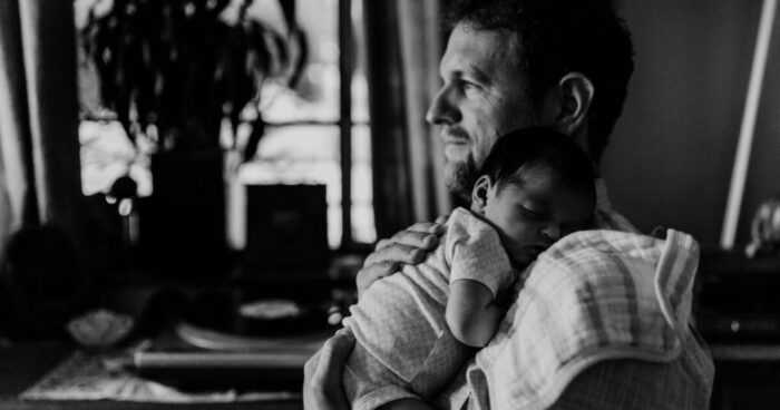 A black and white photo of a man looking out of a window while holding a newborn baby.