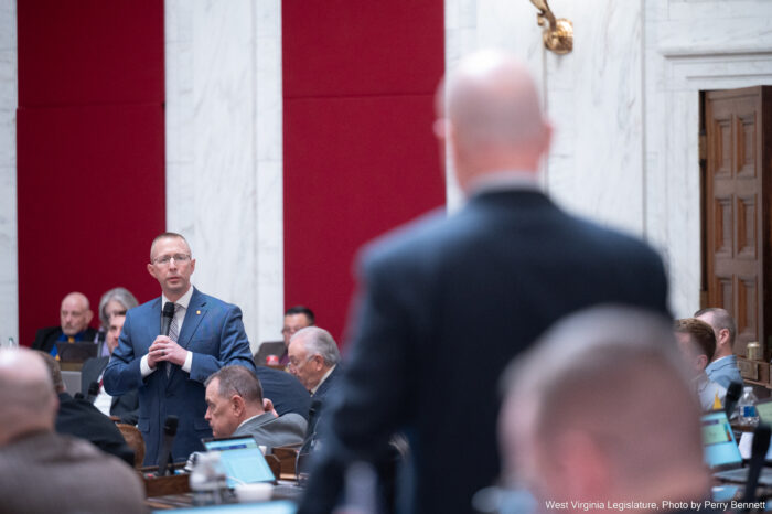 The back of a bald man wearing a suit and facing away from camera is out of focus in foreground. In background but in focus stands a man in a grey blue suit holding a microphone to his chest, appearing to speak. Further behind him are high, white marble walls with red cloth accents running up the wall's lengths. Between the two men, other suited men can be seen sitting at wooden desks and facing left.