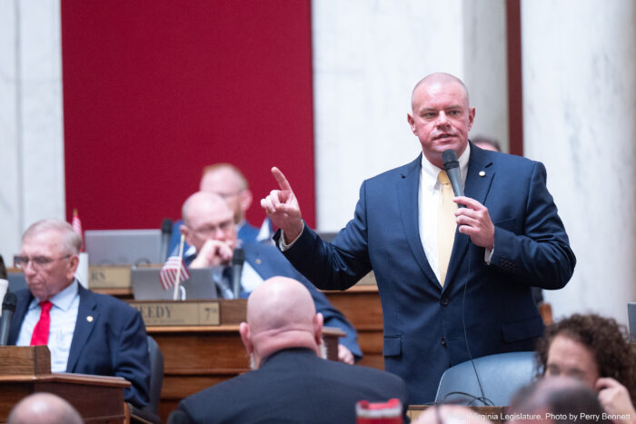 A bald man wearing a blue suit over a gold tie and a white shirt lifts a finger on one hand while holding up a microphone to speak with his other hand. More bald, suited men can be seen behind and in front of him seated at wood desks. In the far background a white marble wall with red cloth accents can be seen.