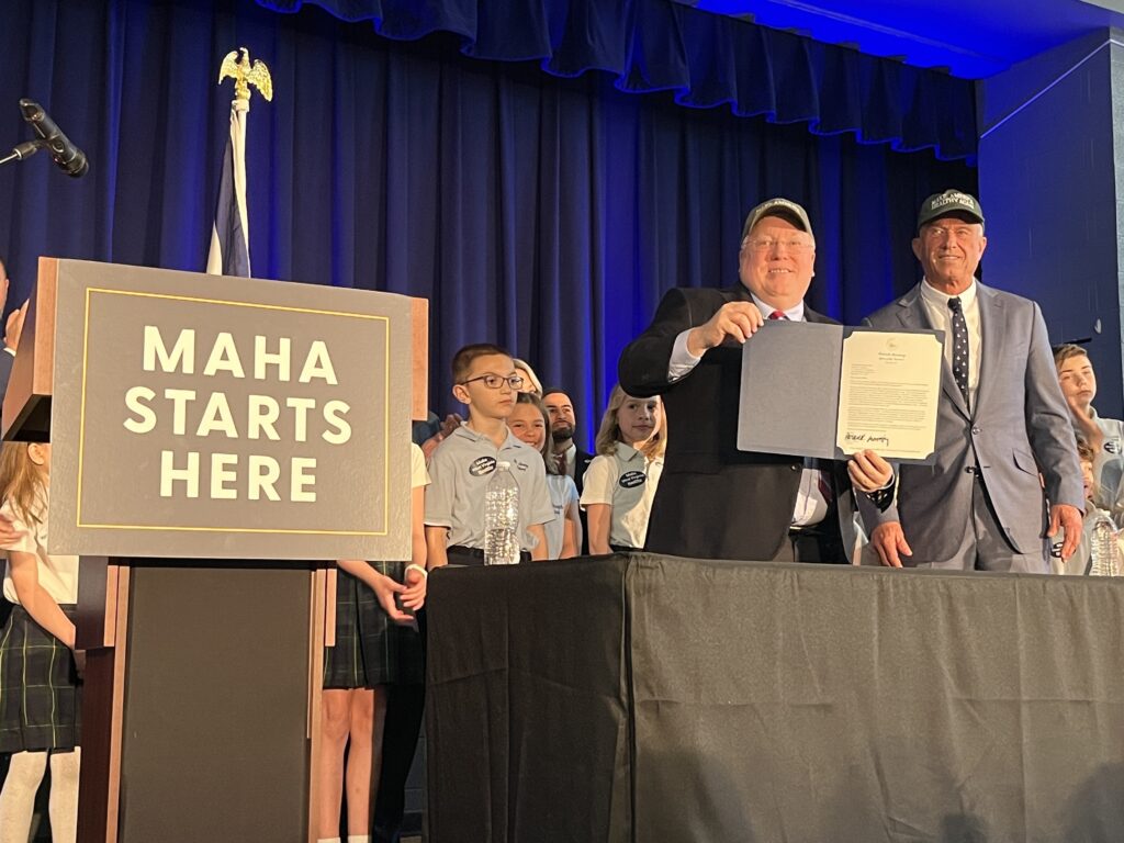 Two men stand at a desk with school children behind them. They both wear hats, and the one on the let holds up a paper folder. To their left, a podium reads "MAHA Starts Here."