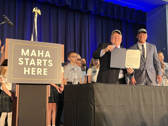 Two men stand at a desk with school children behind them. They both wear hats, and the one on the let holds up a paper folder. To their left, a podium reads "MAHA Starts Here."