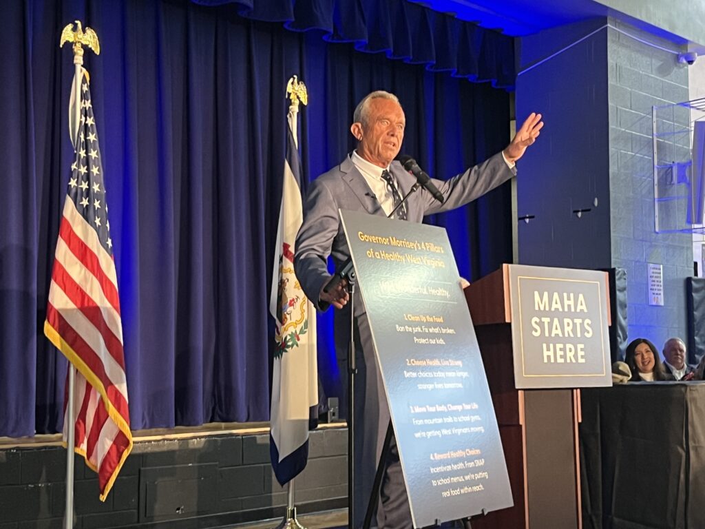 A man in a suit and tie stands at a podium and waves to the audience. Beside him is a sign that reads "Make America Healthy Again." Behind him are blue curtains and the American and West Virginia flags.