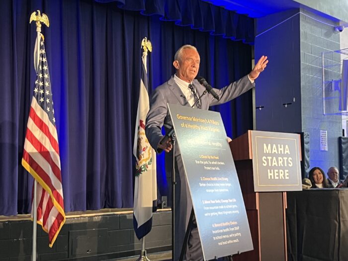 A man in a suit and tie stands at a podium and waves to the audience. Beside him is a sign that reads "Make America Healthy Again." Behind him are blue curtains and the American and West Virginia flags.