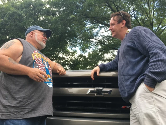 Two men stand next to each other leaning against the front of a vehicle talking.