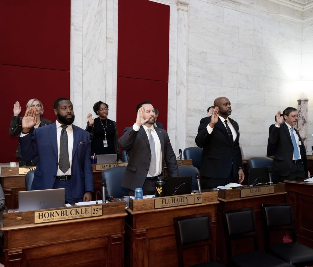 Several people in formal attire stand behind wooden desks with bronze nametags and hold a hand in the air as if saying an oath.