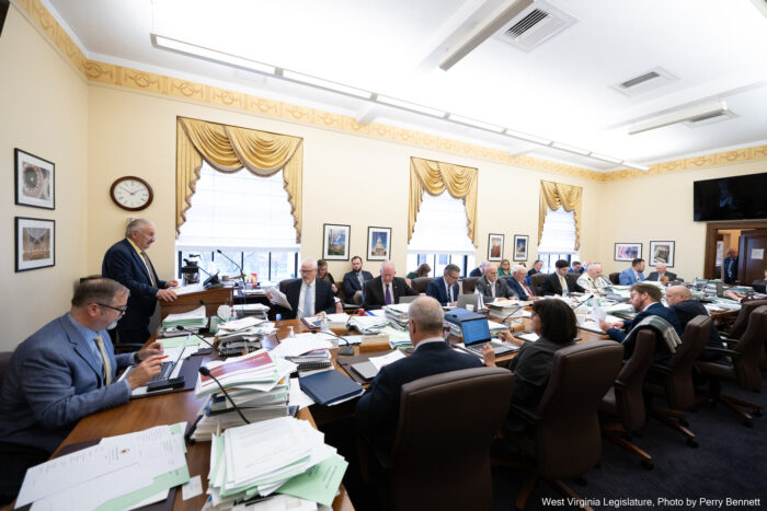 Man stands at a podium in front of a group of others with papers all over desks
