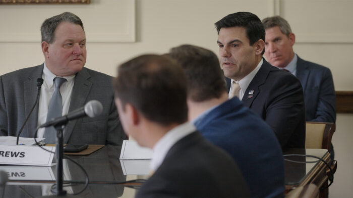 A group of men wearing coats and ties sit at a table and look at a dark-haired man in the corner while he speaks.