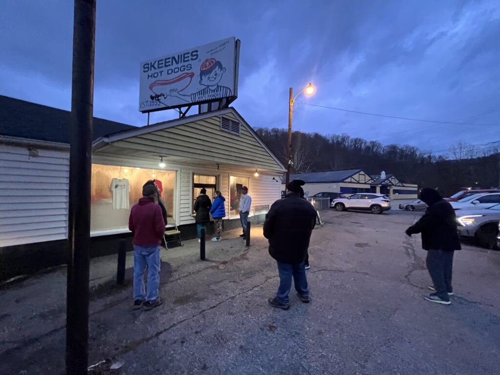 Multiple people standing outside a building with a sign on top that reads, "Skeenies Hot Dogs" in the evening time. There is a cartoon drawing of a little boy holding a hot dog on the sign.