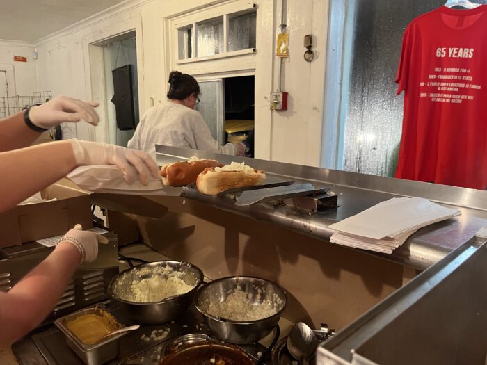 Multiple people standing in a kitchen scooping toppings onto a hotdog. One person standing at a window serving customers.