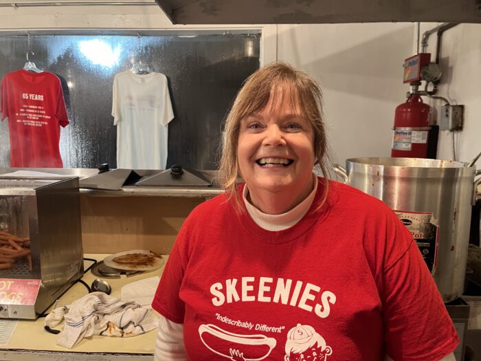 White woman with blonde hair wearing a t-shirt that says "Skeenies" with an image of a hot dog on the t-shirt.