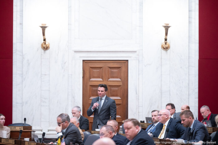 A man stands up and speaks to into a microphone. He is surrounded by other men in suits.