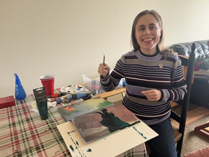 A woman in a purple striped sweater sits at a kitchen table, with a storybook covered in green paint before her. She holds up a paintbrush and smiles toward the camera.
