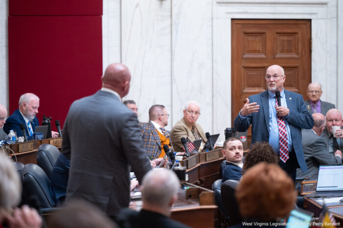 Dozens of people sit at wooden desks dressed in formal attire. Two stand up and hold microphones, facing each other as if having a conversation. The room has white marble walls with areas covered in red velvet. It is the floor of the West Virginia House of Delegates.