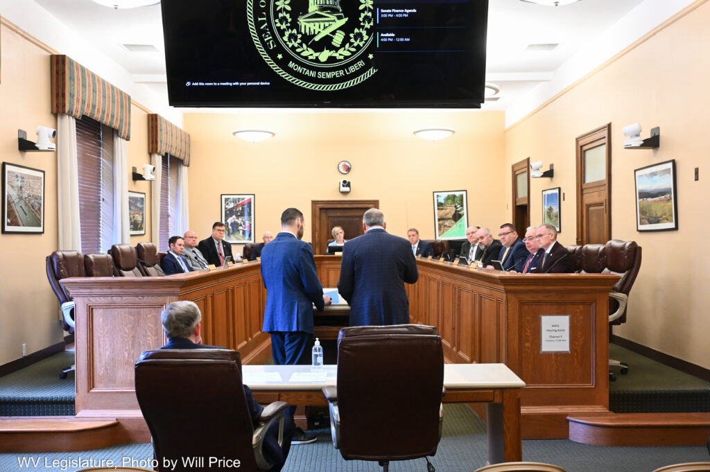 A black screen hangs over two men standing at the head of a u-shaped wooden dais desk. Behind the standing men is a man seated in a large leather office chair. Seated at the desk are many suited men.