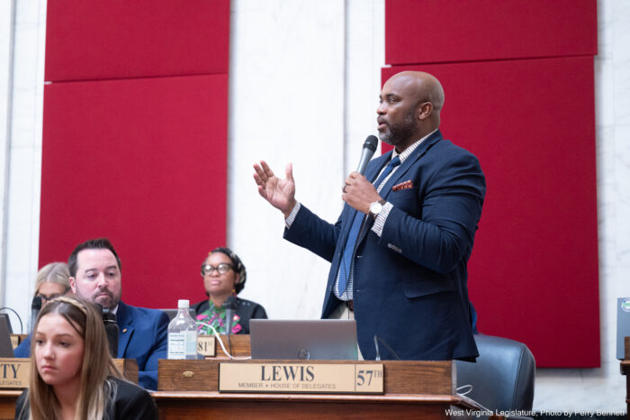 A man wearing a black suit and blue tie with red pocket square speaks on the floor of a legislative body. He speaks into a microphone, gesturing with his right hand.