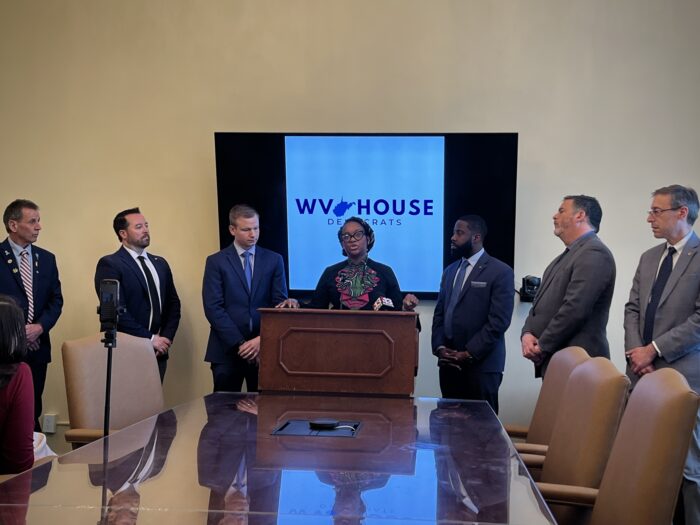 People dressed in formal attire stand at the front of a room before a long, wooden table. They are centered around a podium, where one women speaks into a microphone and gestures with her left hand. Behind them, a screen reads "WV House Democrats."