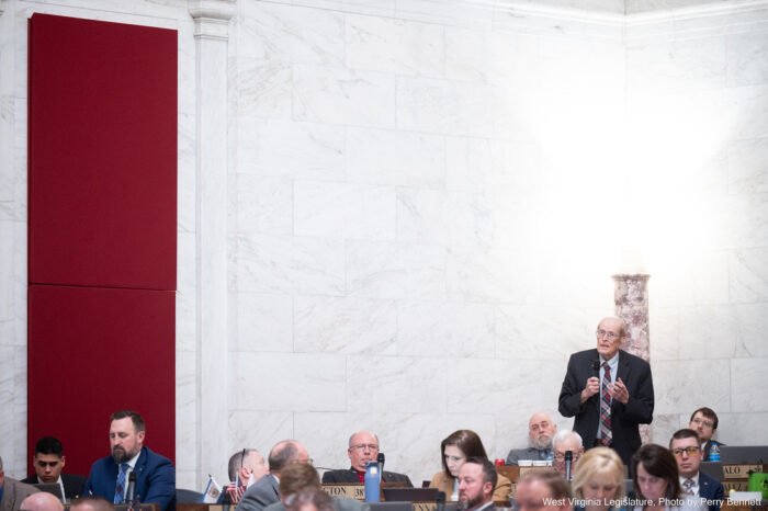 A man in a suit stands at a wooden desk and speaks into a microphone. He is surrounded by others sitting at desks in the House chamber.