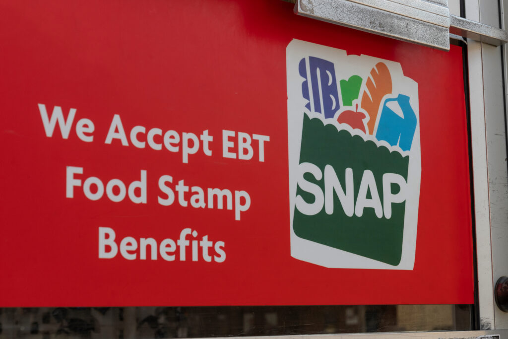 A red sign covers a glass door with a push handle. The sign says, "We accept EBT Food Stamp Benefits" in white font with a graphic of food in a green basket to the right.