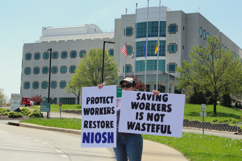A woman holds two signs that read "Protect Workers Restore NIOSH" and "Saving Workers is not Wasteful" in front of a white concrete building punctuated by circular, blue-glass windows on a sunny day